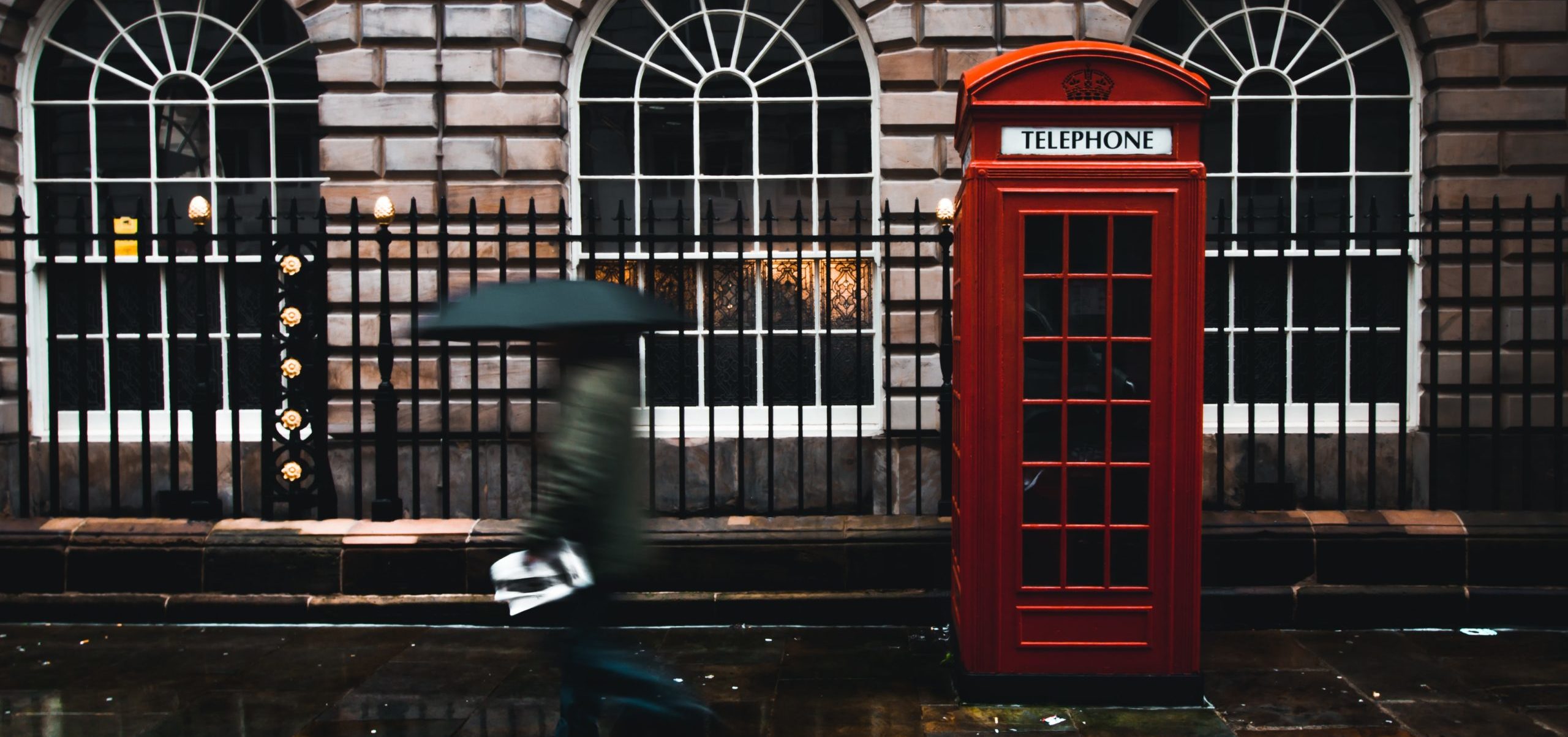 A red, British telephone box on a rainy sidewalk, with large arched windows behind it and a man passing quickly carrying an umbrella.